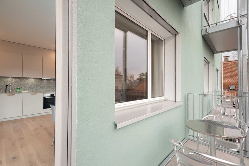 The image shows a modern apartment interior with a balcony or terrace. The room has large windows that provide a view of the exterior, and the walls are painted in a light mint green color. The kitchen can be seen in the background, with white cabinets an