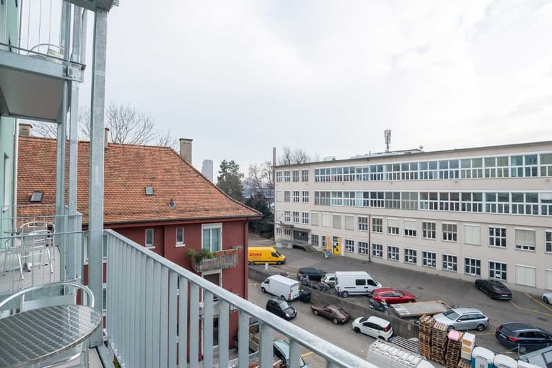 The image shows a multi-story commercial or office building with a parking lot in the foreground. The building has a mix of modern and older architectural styles, with a red-tiled roof on one side and a more contemporary glass and steel facade on the othe