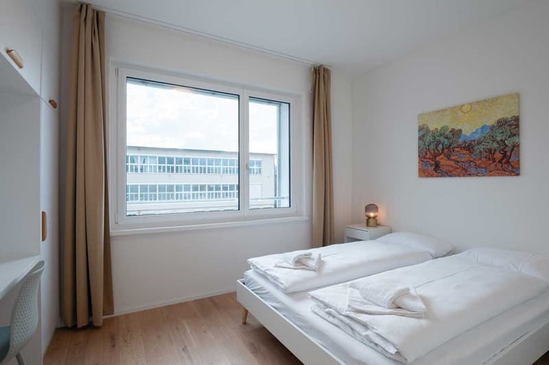 Spacious bedroom with large windows providing a view, white walls, hardwood floors, a bed with white bedding, and a decorative painting on the wall.
