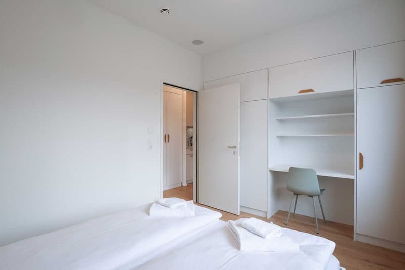 Spacious bedroom with white walls, hardwood floors, large closet, desk, and a balcony or terrace visible through the open door.