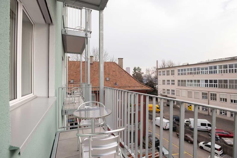 Balcony with metal railing, outdoor furniture, overlooking a street with buildings and parked cars