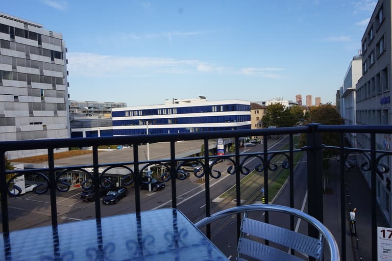 The image shows a multi-story office building with a balcony or terrace overlooking a street with parked cars. The building has a modern, glass and steel design with a blue and white color scheme. The surrounding area appears to be an urban setting with o