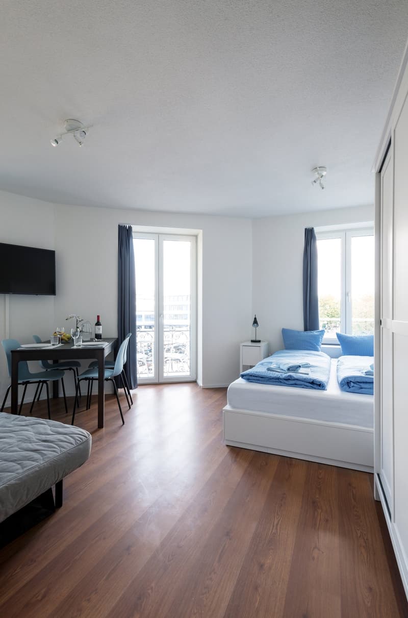 The image shows a studio apartment with a living and sleeping area. The room has hardwood floors, white walls, and large windows that provide natural light. The sleeping area features a white bed with blue bedding, and the living area has a dining table w