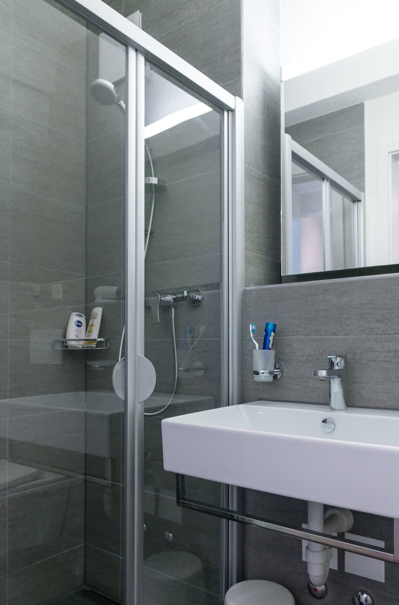 Modern bathroom with gray tile walls, glass shower enclosure, white sink, and bathroom accessories