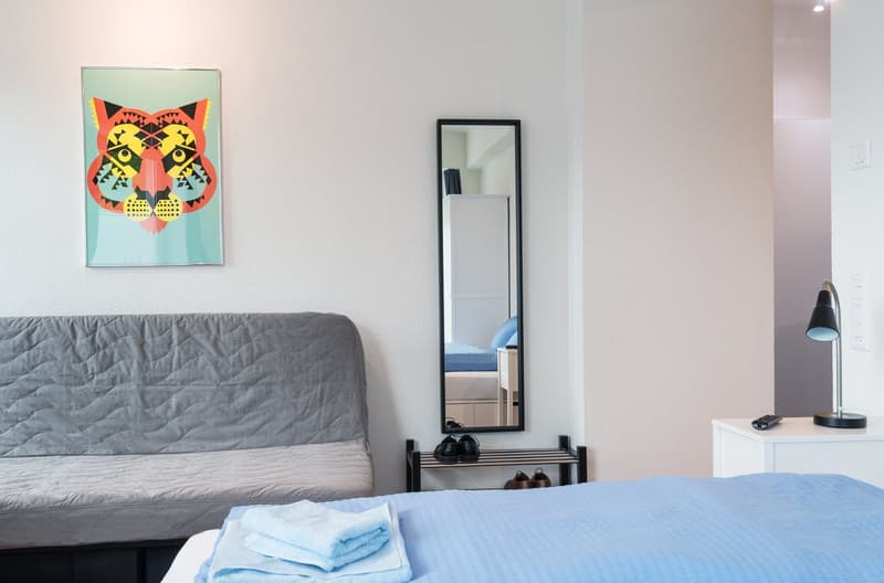 Bedroom with a gray sofa, a full-length mirror, a shoe rack, and a colorful abstract artwork on the wall. The room also has a lamp and a blue bedding set.