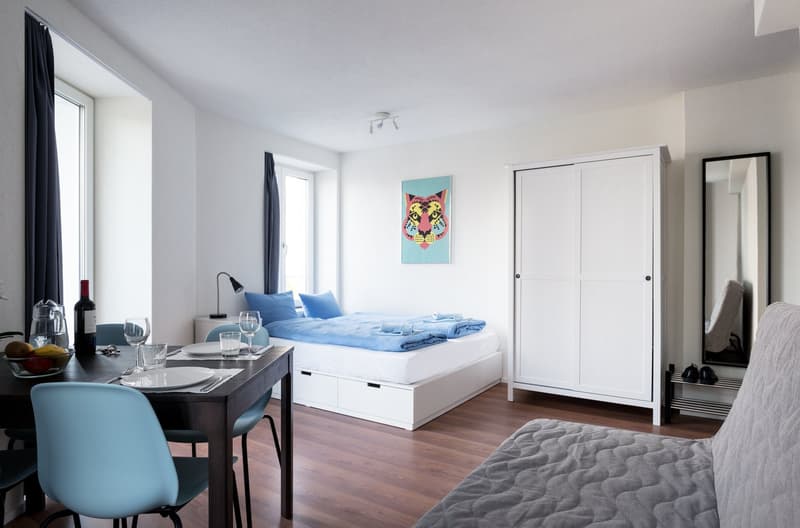 Spacious studio apartment with hardwood floors, white walls, large windows, a bed with blue bedding, a dining table, and a kitchenette with a dishwasher. The room is decorated with a colorful wall art piece.