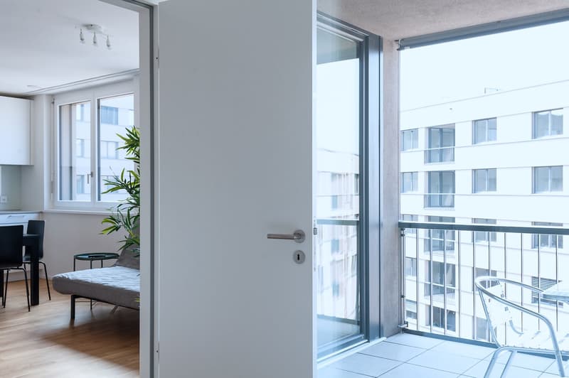 Open floor plan with living room, bedroom, and balcony. Large windows providing natural light and a view of the surrounding buildings.