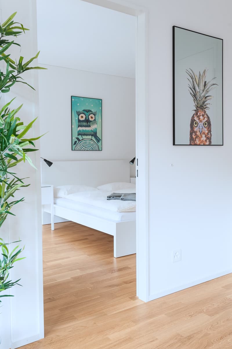 White walls, hardwood floors, framed artwork featuring an owl and a pineapple, potted plants, and a bed visible through the open doorway
