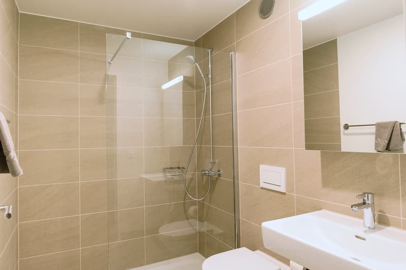 Spacious bathroom with beige tile walls, glass shower enclosure, sink, and towel rack.