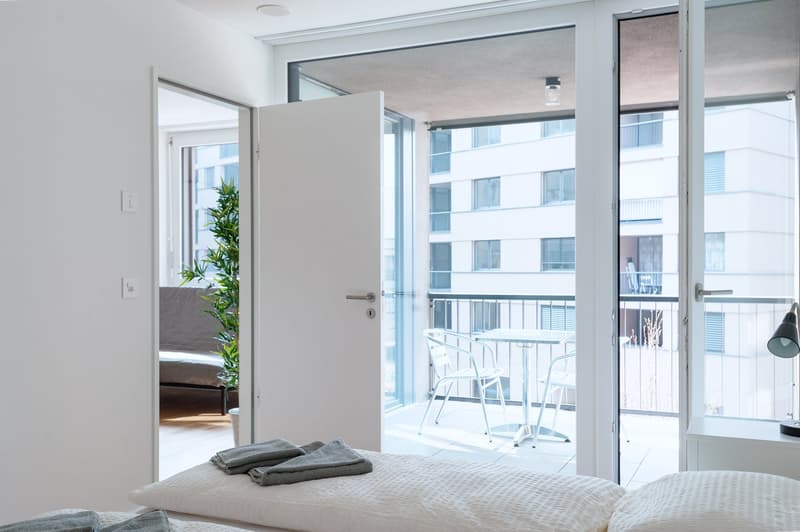 Spacious bedroom with large windows, sliding glass door leading to a balcony, white bedding, and a potted plant.