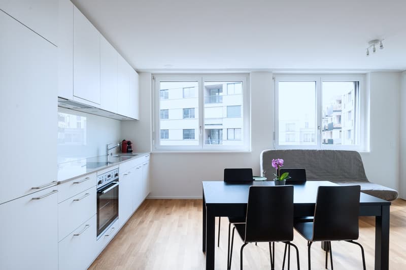 Modern, open-concept kitchen and dining area with white cabinets, stainless steel appliances including a dishwasher, and large windows overlooking the city. The dining table has four black chairs and there is a gray sofa in the corner.