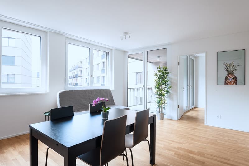 Spacious open-plan living area with large windows, hardwood floors, black dining table and chairs, gray sofa, potted plant, and artwork on the wall. The apartment appears to have access to a balcony or terrace.