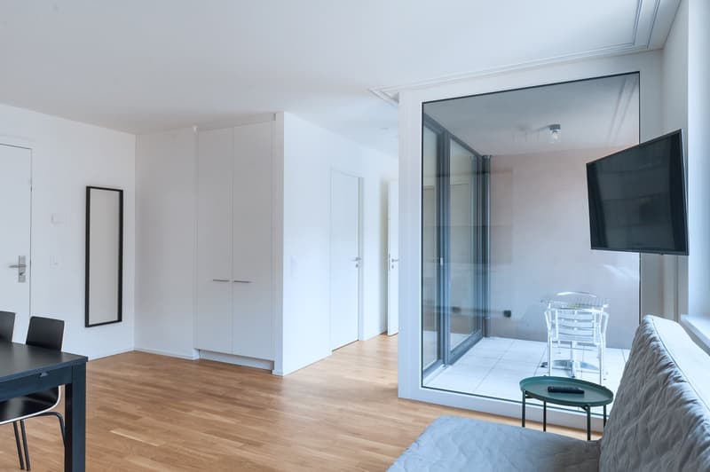 Spacious open-plan living area with hardwood floors, white walls, and large sliding glass doors leading to a balcony or terrace. The room features a sofa, coffee table, and a mounted TV. There are also built-in wardrobes and a desk area visible.