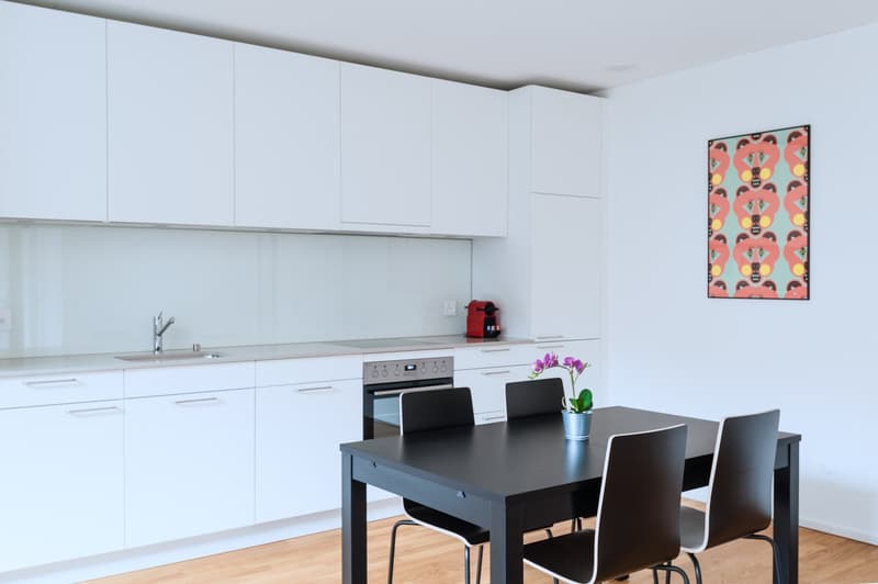 Modern white kitchen with black dining table and chairs, stainless steel appliances, and a colorful abstract artwork on the wall