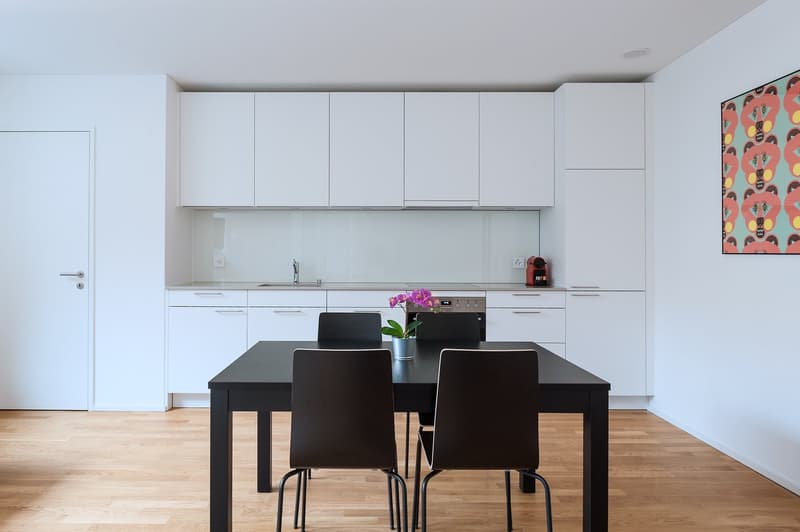 Modern, minimalist kitchen with white cabinets, black countertop, dining table, and colorful artwork on the wall