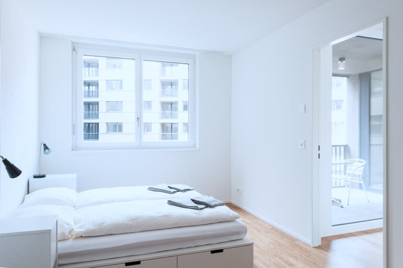 Spacious bedroom with large windows, hardwood floors, white walls, and a bed with bedding. There is a balcony or terrace visible through the windows.