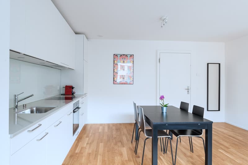 Open-concept kitchen and dining area with white walls, hardwood floors, modern kitchen appliances including a dishwasher, a dining table with black chairs, and a framed artwork on the wall.