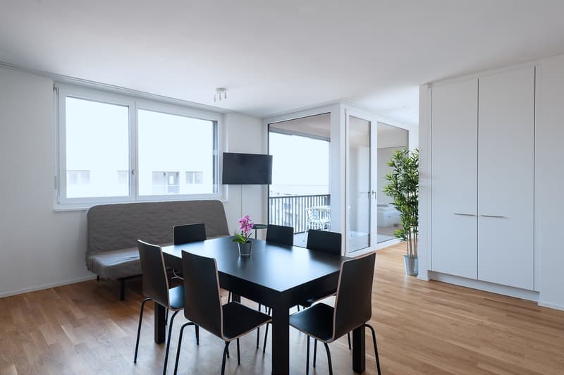 Spacious open-plan living area with large windows, hardwood floors, black dining table and chairs, gray sofa, TV mounted on the wall, and a balcony or terrace visible through the windows.