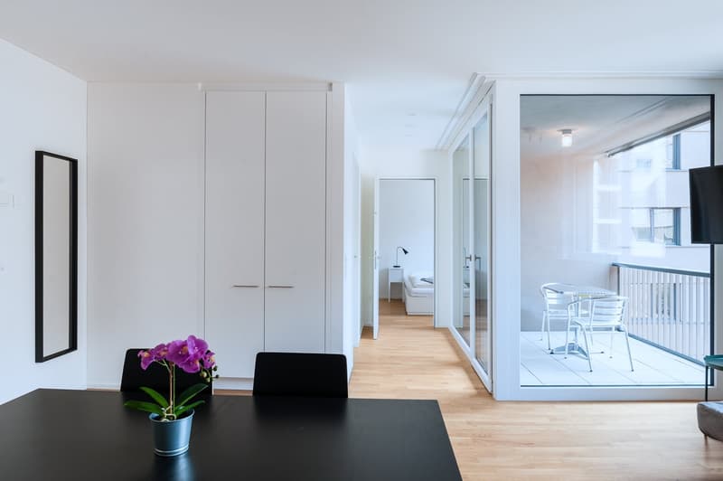 Spacious open-plan living area with white walls, hardwood floors, and large sliding glass doors leading to a balcony. The room features a dining table, chairs, and a potted orchid plant. The hallway in the background suggests additional rooms, including a