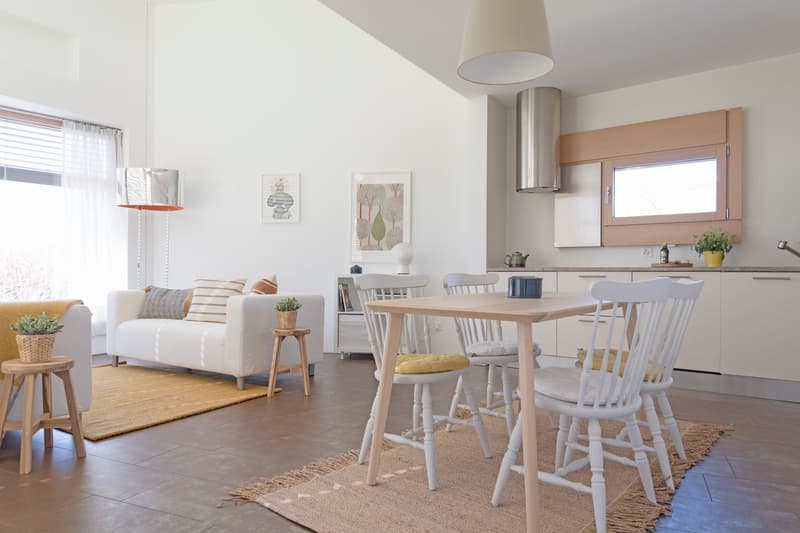 Open-plan living and dining area with white walls, wooden floor, white furniture, and a rug. The living area has a white couch, a side table, and a lamp. The dining area has a wooden table and white chairs. The kitchen has white cabinets, a countertop, an
