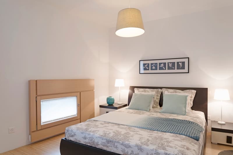 Bedroom with bed, nightstands, lamps, picture frame, wooden headboard, wooden floor, white walls, power outlet, and small window.