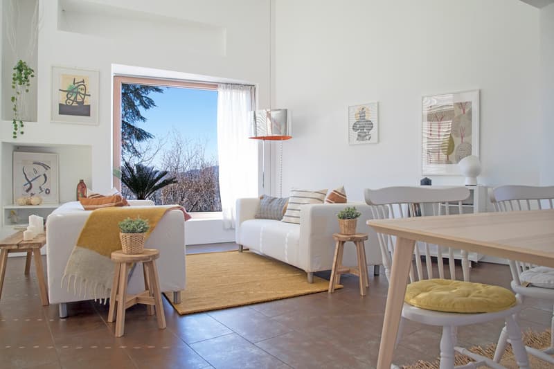 modern living room with white walls, wooden floor, two white sofas, beige rug, wooden stools, dining table, white chairs, large window with white curtains, plants, artwork