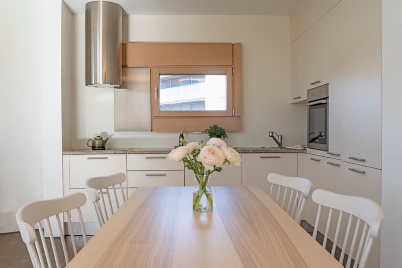 modern kitchen with dining area, wooden table, white chairs, white cabinets, stainless steel appliances, window with blinds, flower vase, teapot, bottle