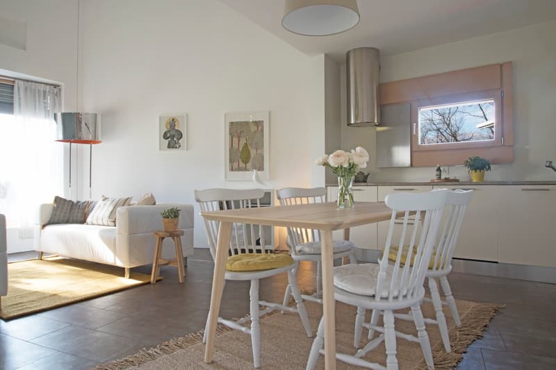 Dining area with a wooden table and white chairs, a white couch with pillows, a rug, a lamp, and a flower vase on the table. The kitchen has a window, a potted plant, and a bottle on the counter.