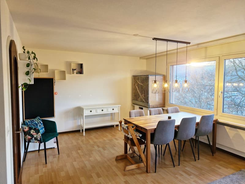 Dining room with wooden table, chairs, white sideboard, blackboard, radiator, hanging lamps, plants, and a view of trees outside