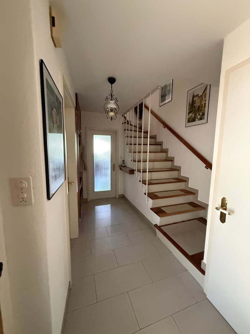wide hallway with white tile flooring, a staircase with wooden steps and white railings, and a white door on the right. There are also some picture frames hanging on the wall.