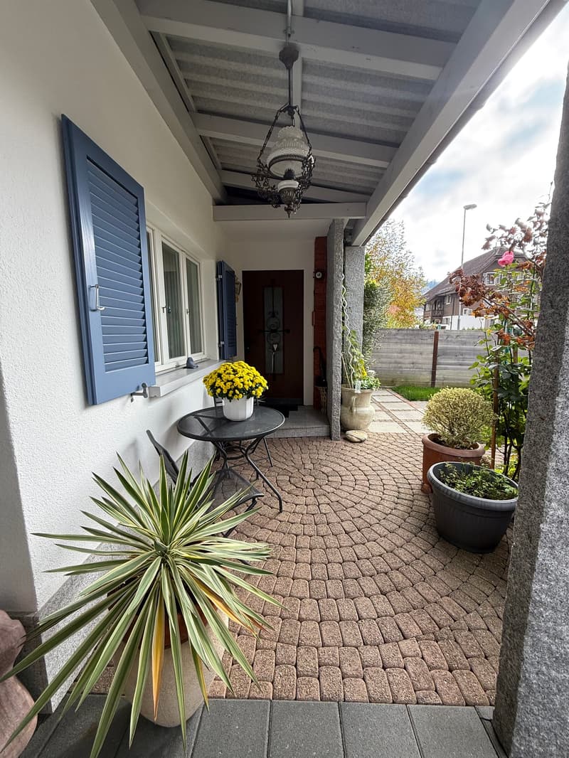covered patio with brick flooring, blue window shutters, outdoor table and chairs, hanging light fixture, various potted plants