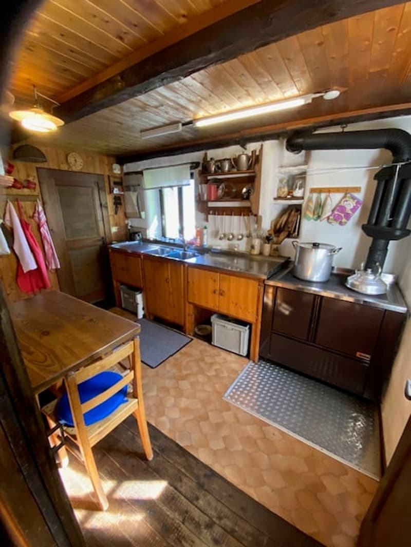 wooden cabinets, stove with chimney, sink, table and chair, hanging pots and pans, shelves with various items