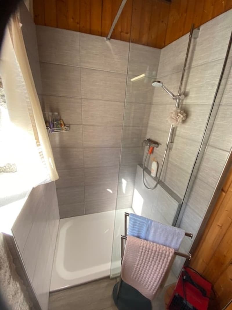 Shower with showerhead, tiles, shower gel dispenser, towels on towel rack, towels on chair, window with curtains, wooden walls, floor mat
