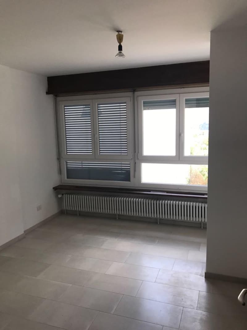 An empty room with tile flooring, white walls, and a ceiling light fixture. Below the windows, there is a wooden bench with a radiator below it.