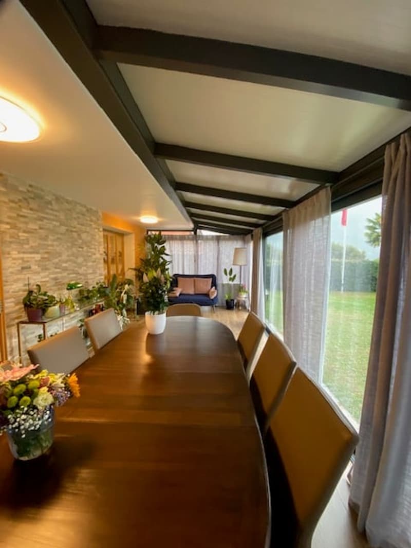 Long dining table with chairs, flower vase, ceiling lamp, stone wall, glass windows, sofa with cushions, potted plants, garden view