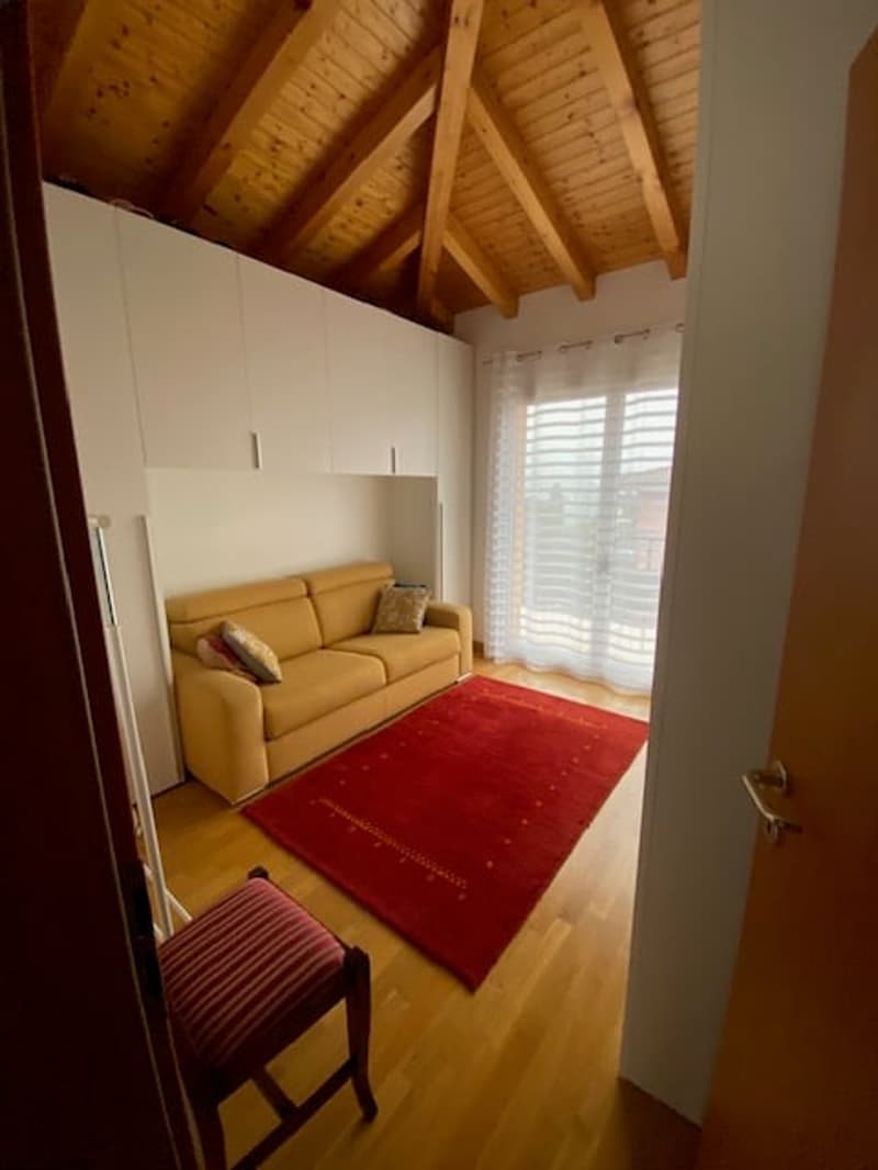 Living room with wooden floor, beige sofa, red rug, wooden chair, white cabinets, wooden beams, large window with white curtain