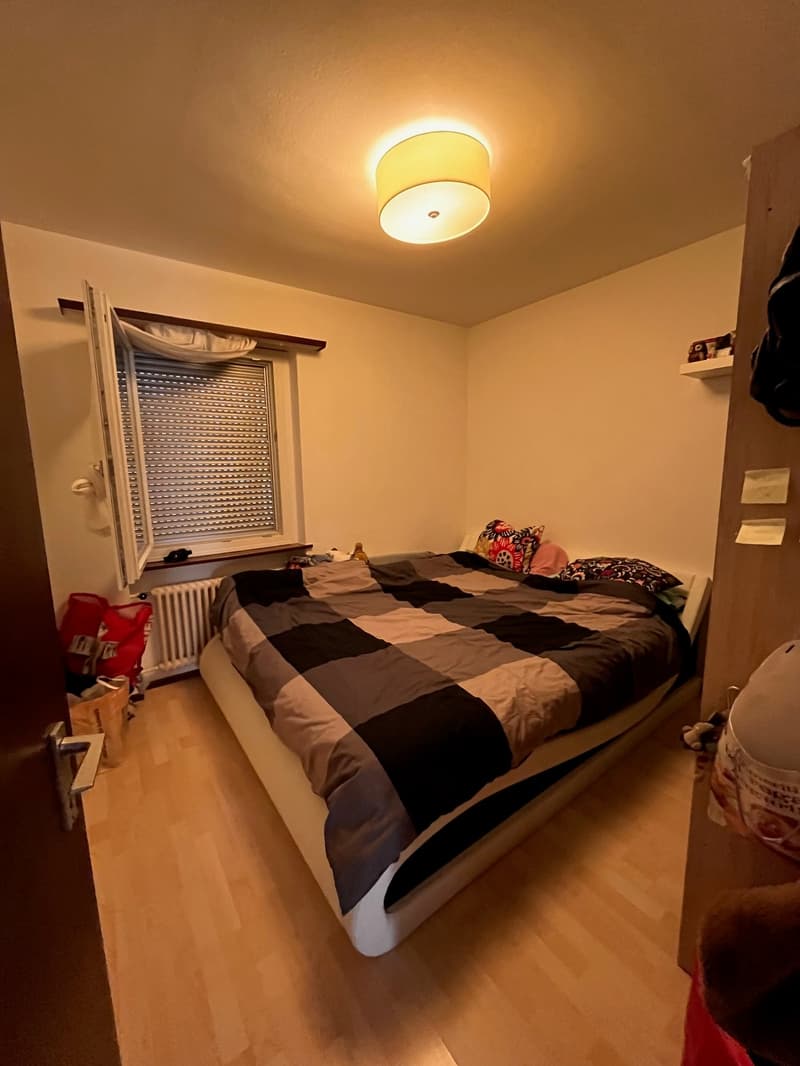A bedroom with wooden flooring and white walls. The room has a window with blinds and a radiator below. It features a bed with a plaid sheet, surrounded by pillows. There are bags and other items on the floor near the bed.
