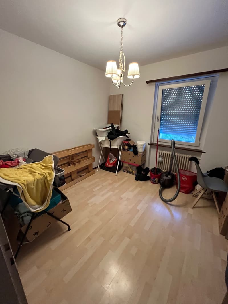 Empty room with wooden flooring, a chandelier, window with blinds, wooden pallets, storage boxes, a radiator, a vacuum cleaner, and other cleaning supplies.