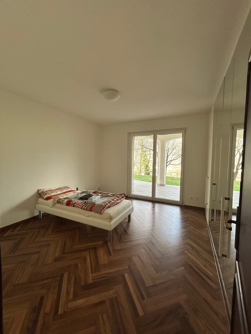 Spacious bedroom with large windows overlooking a green outdoor area, hardwood flooring, and a bed with a patterned comforter.
