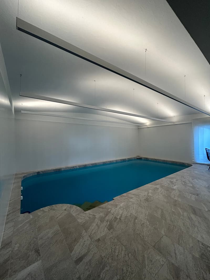 The image shows an indoor swimming pool with a large, rectangular pool filled with blue water. The pool is surrounded by a gray, textured tile floor. The ceiling has recessed lighting and a long, horizontal beam running across the length of the room.