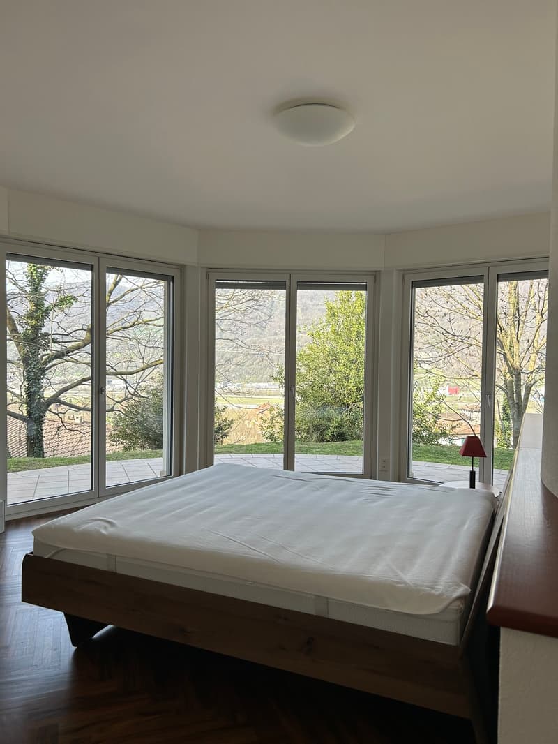 Spacious bedroom with large windows offering a scenic view of the outdoor greenery. The room features a white bed and hardwood flooring.