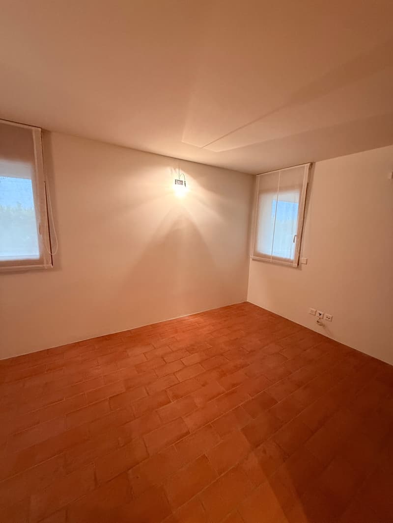 Empty room with white walls, brown tile floor, and two windows with white curtains. There are power outlets on the wall.