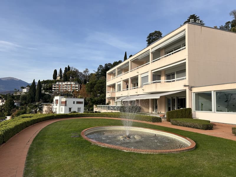 Modern apartment building with balconies, surrounded by greenery and a fountain, with a scenic view of mountains