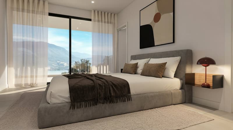 A bedroom with a bed, a nightstand, a lamp, a rug, and a large window with a view of the mountains and the sea.