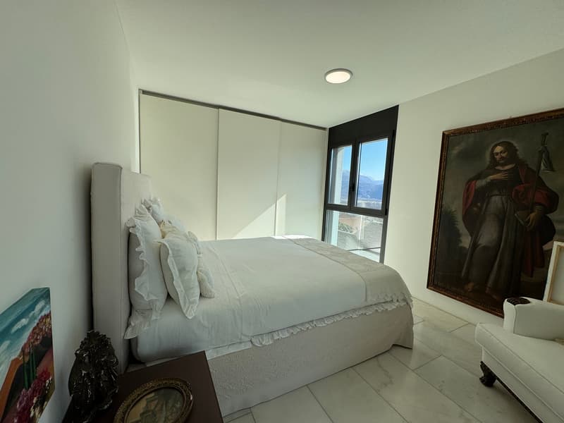 Master bedroom with white bedding and pillows, large white walls, bedside tables, artwork, sliding window with a view of the mountains