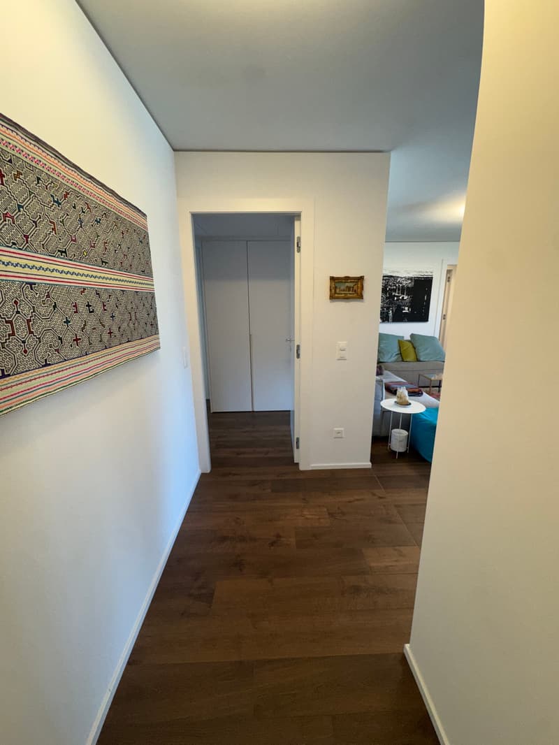 Empty hallway with wooden floor, white walls, and a rug on the left wall. A white door leads to a living room with a couch, table, and picture frame on the wall.
