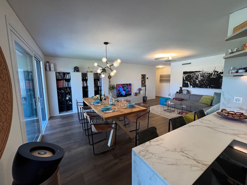 Dining room with wooden table, chairs, chandelier, TV, bookshelves, couch, rug, kitchen counter, speaker