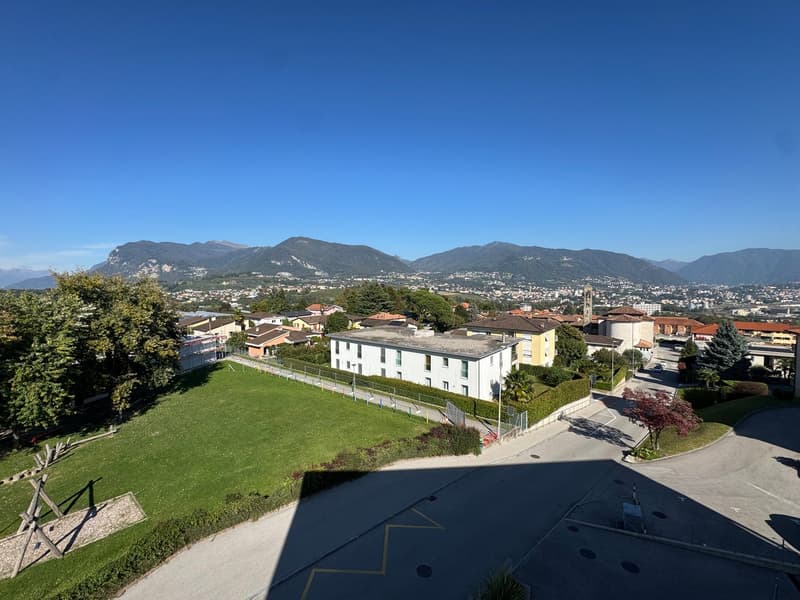 Aerial view of a residential area with a large grassy field, a white building, and a road with a parking lot. Mountains are visible in the distance.