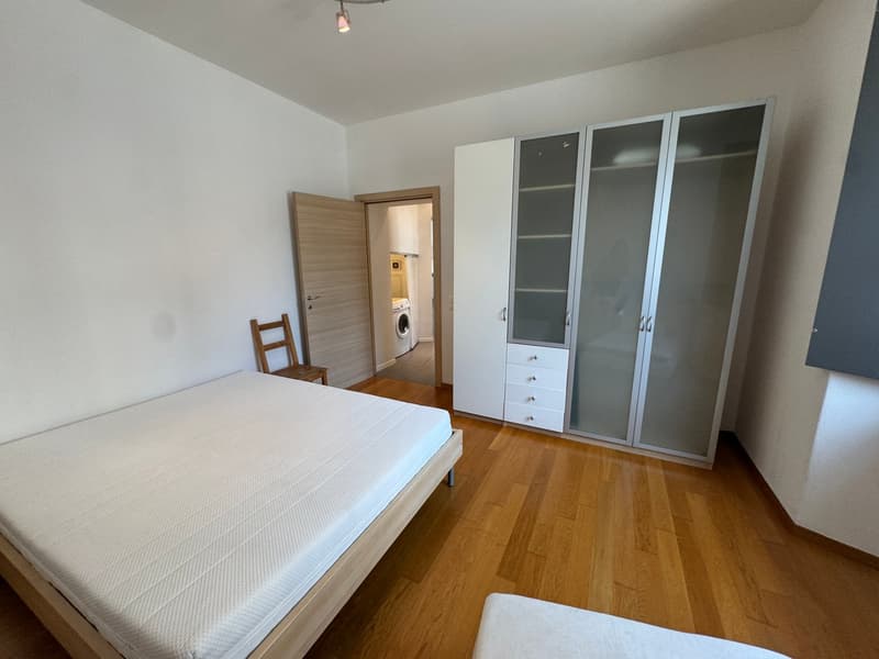 A bedroom with a bed, wooden floor, white walls, a wooden chair, a wardrobe with glass doors, and a washing machine in the hallway.