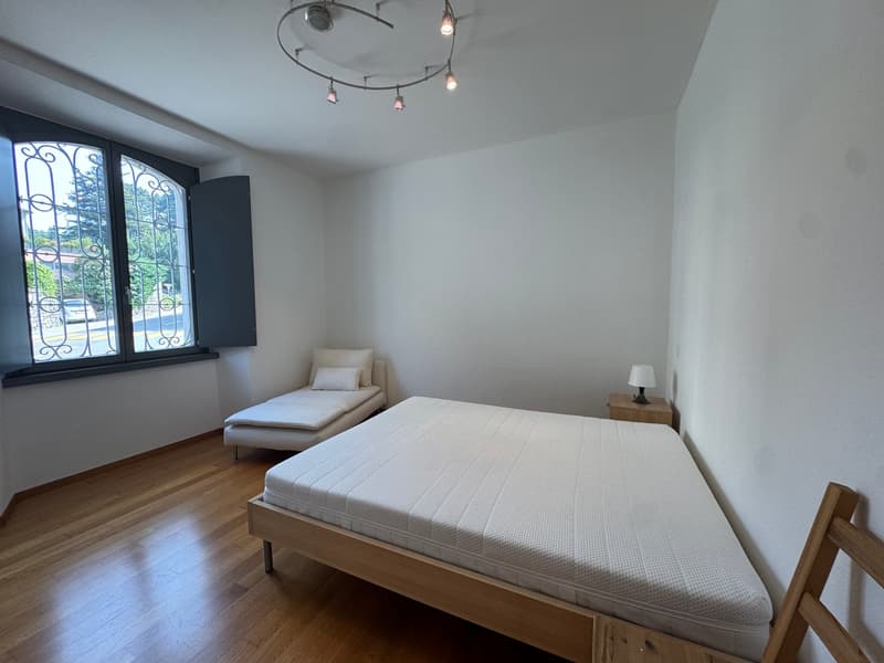 A bedroom with a double bed, a single bed, a nightstand, a lamp, a window with a view of the outside, and a wooden floor.